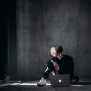 Young man sitting on a concrete floor using a laptop. natural light comes in from a window on the left.